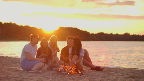 Two-men-and-three-young-girls-are-sitting-around-bonfire-on-the-beach-with-beer.-One-of-the-girls-is-checking-her-mobile-phone-and-turning-music-at-sunset-in-summer-evening-on-the-lake-coast.